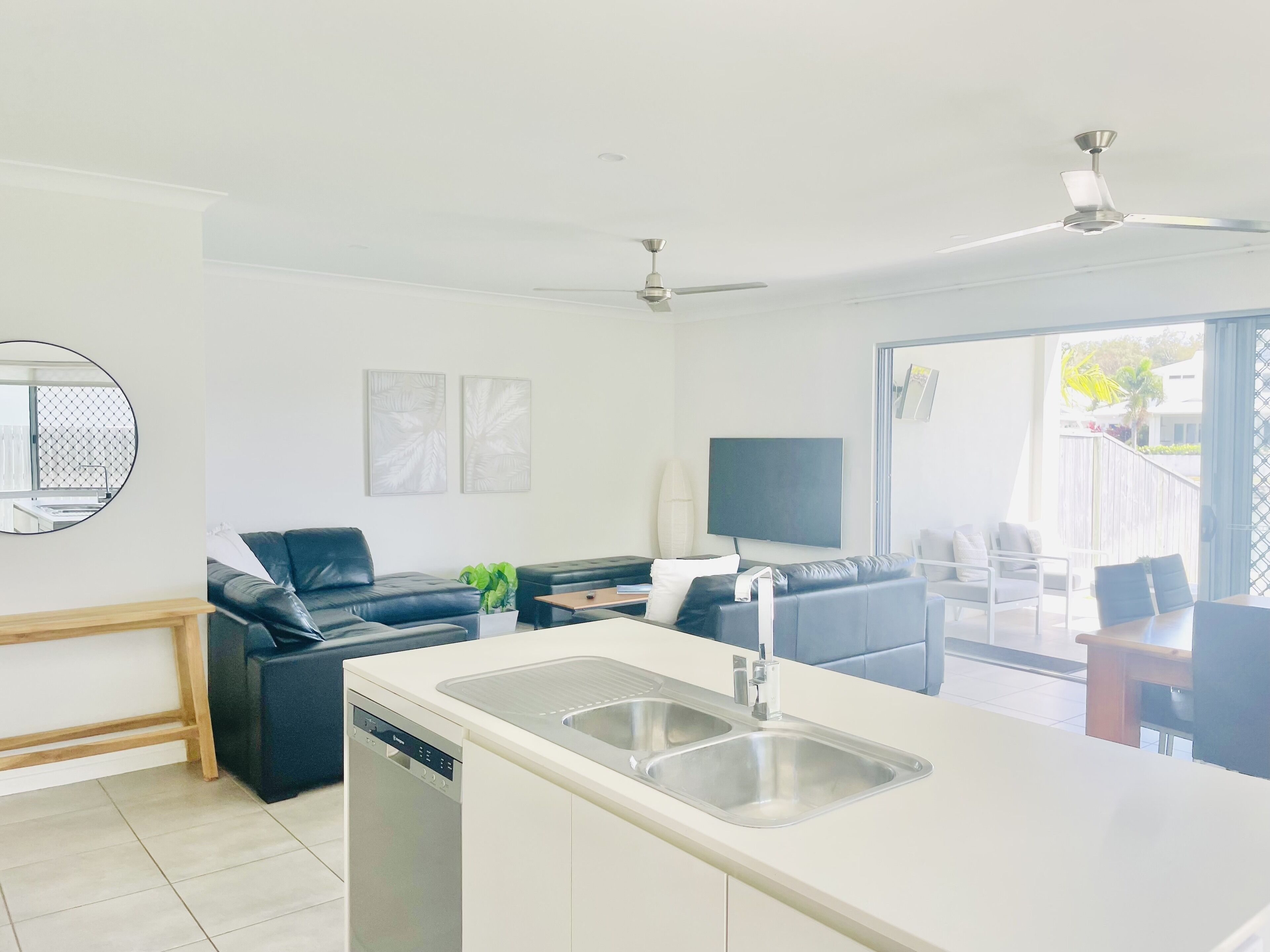 A modern kitchen with a dishwasher, a dining area with a table and chairs, a black leather sofa, and a flat-screen TV mounted on the wall.