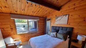 Un dormitorio acogedor de cabaña de madera con una cama grande, una mesita de noche, una silla y una ventana con vistas al exterior.