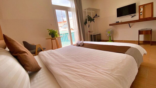 A hotel room with a large bed, a chair, a desk, a TV, and a balcony with a view of buildings. - HANZ Chuong Hotel Da Lat (Xuan Huong - Da Lat)