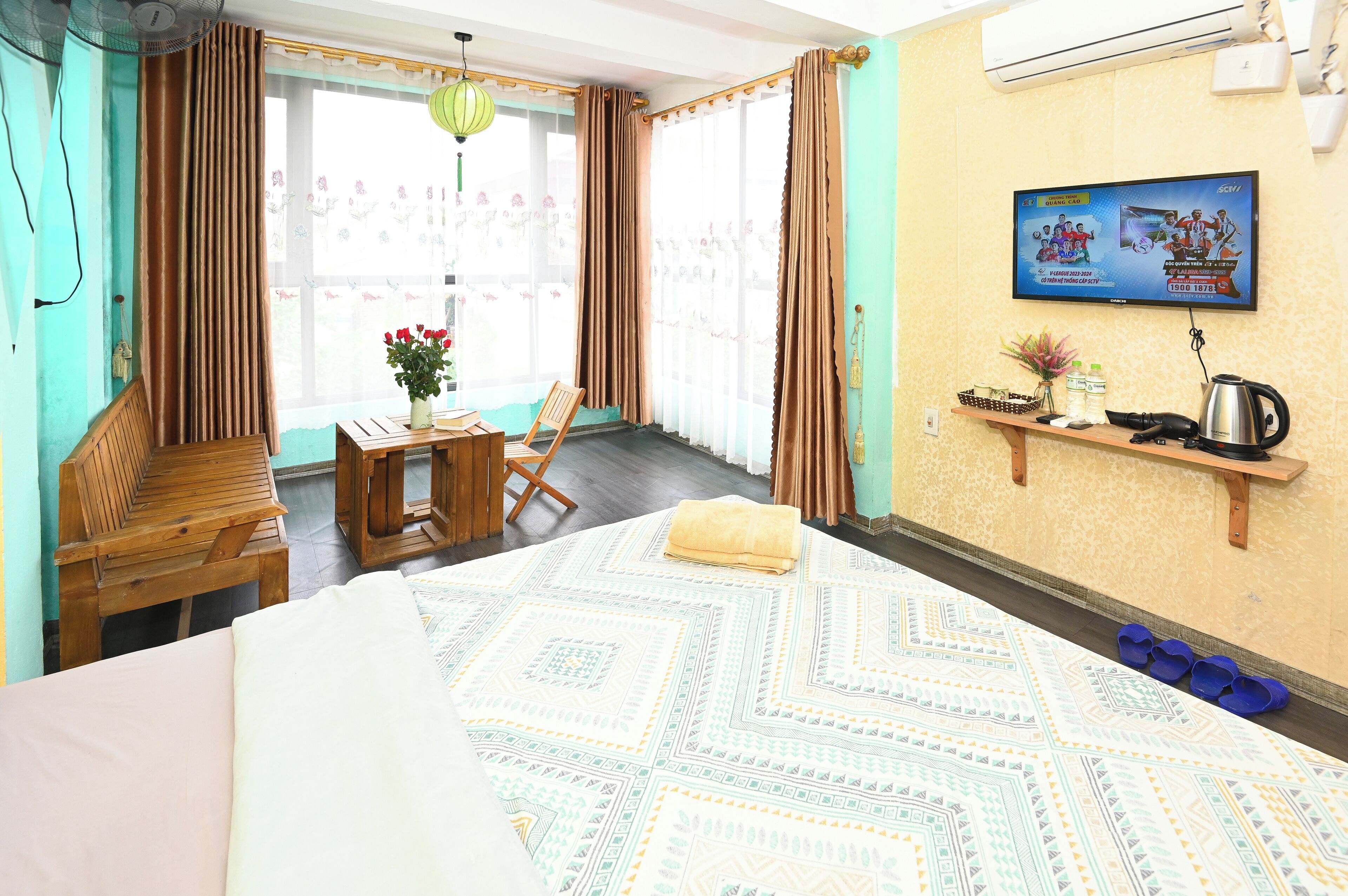 Une chambre d’hôtel avec un lit, une chaise en bois, une table ornée d’un vase de fleurs, un téléviseur fixé au mur et une étagère avec divers objets.