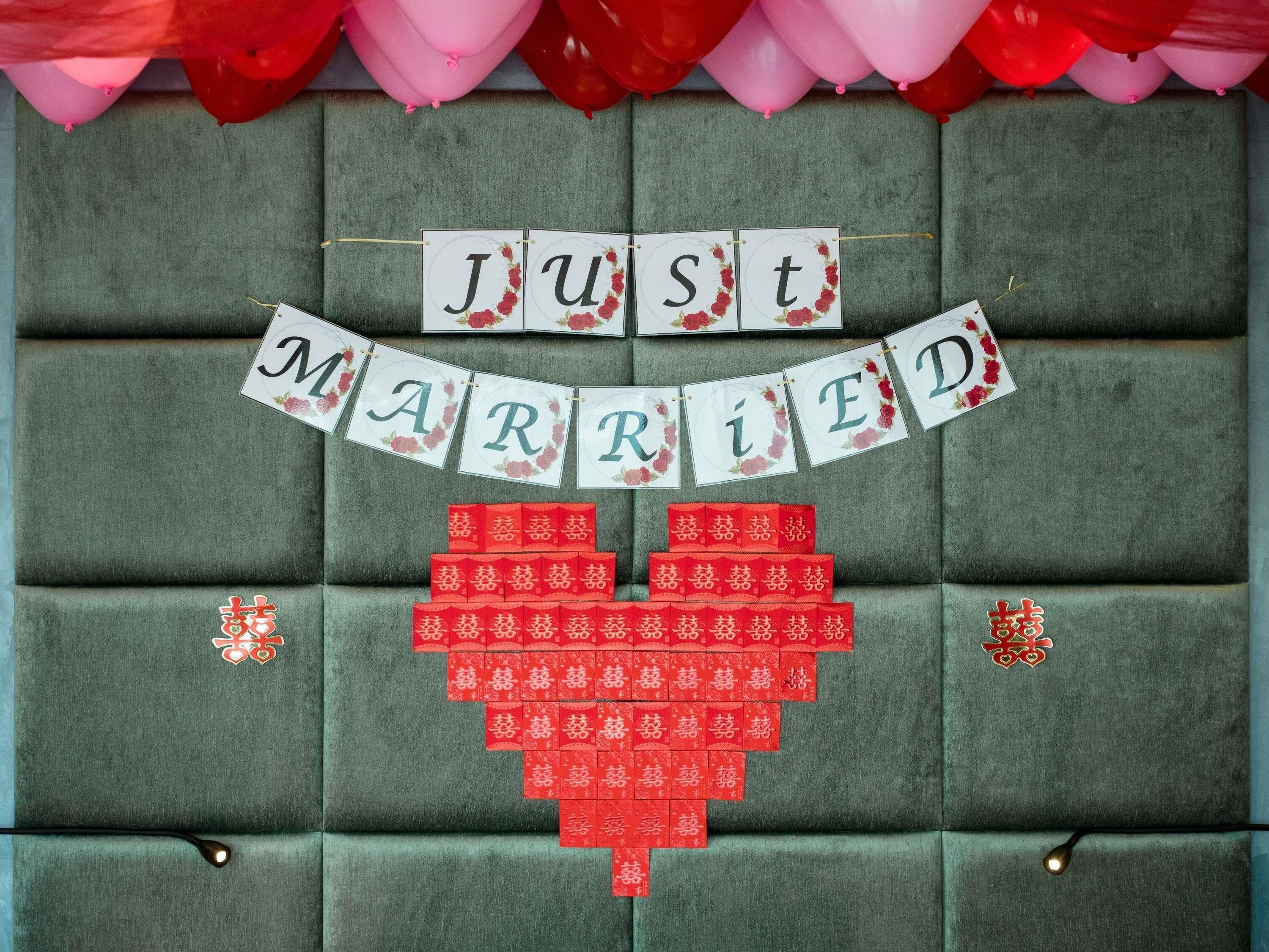 A room with a padded headboard, a banner reading 'JUST MARRIED,' and red envelopes arranged in a heart shape.