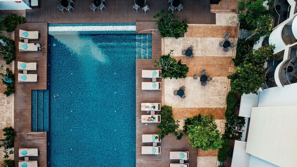 Vista aérea de un hotel con una amplia piscina, varias zonas de descanso con sillas y mesas, y abundante vegetación circundante.