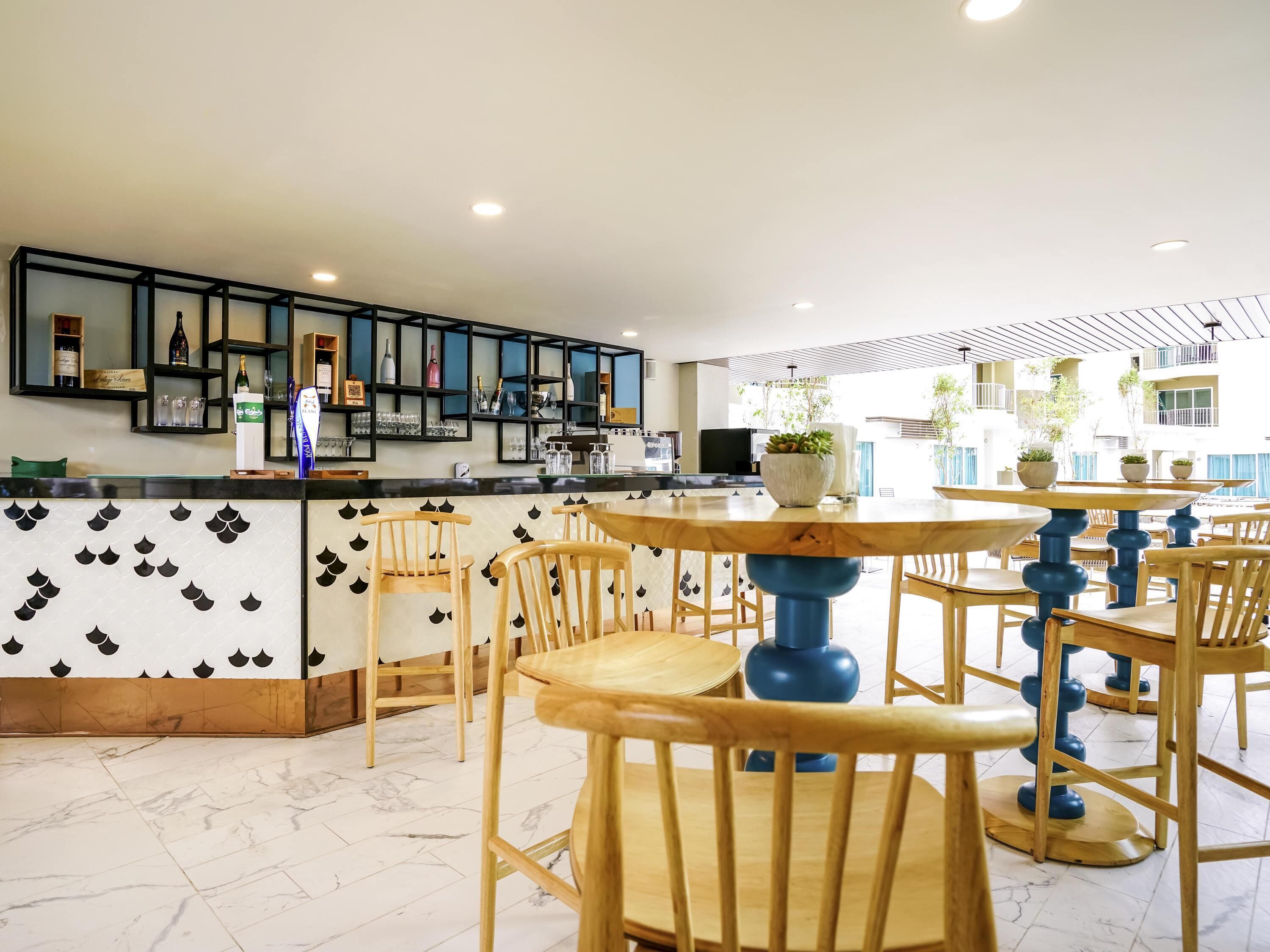A modern bar area with wooden chairs and tables, a counter with various bottles, and a tiled floor.