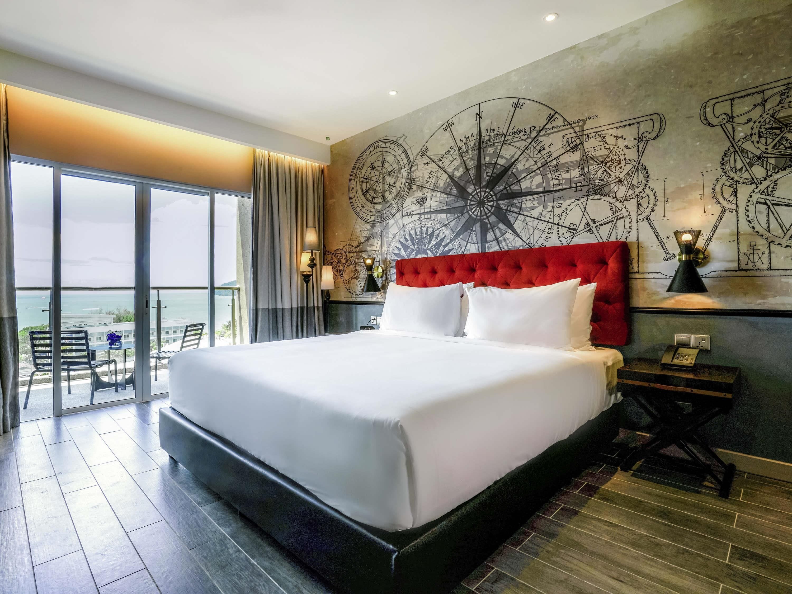 A large bed with a red headboard, a wall clock, and a large window with a view of the sea.
