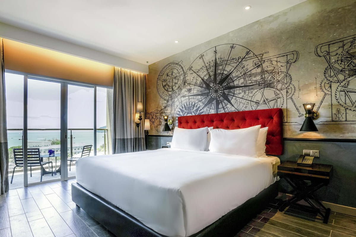 A large bed with a red headboard, a wall clock, and a large window with a view of the sea.