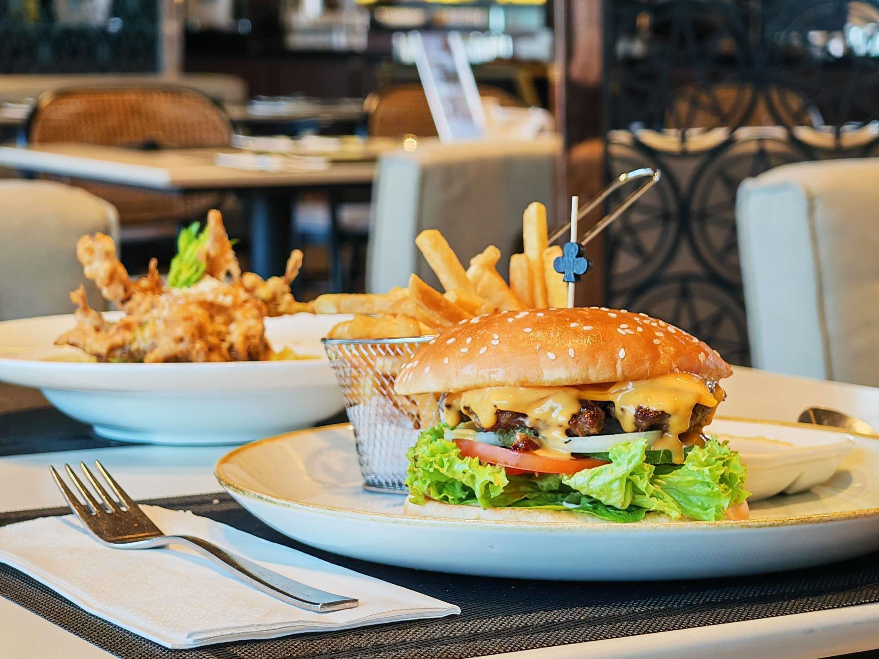 A restaurant setting with a table set for two, including plates with a burger and fries, a basket of fries, and a fork and knife on a napkin.