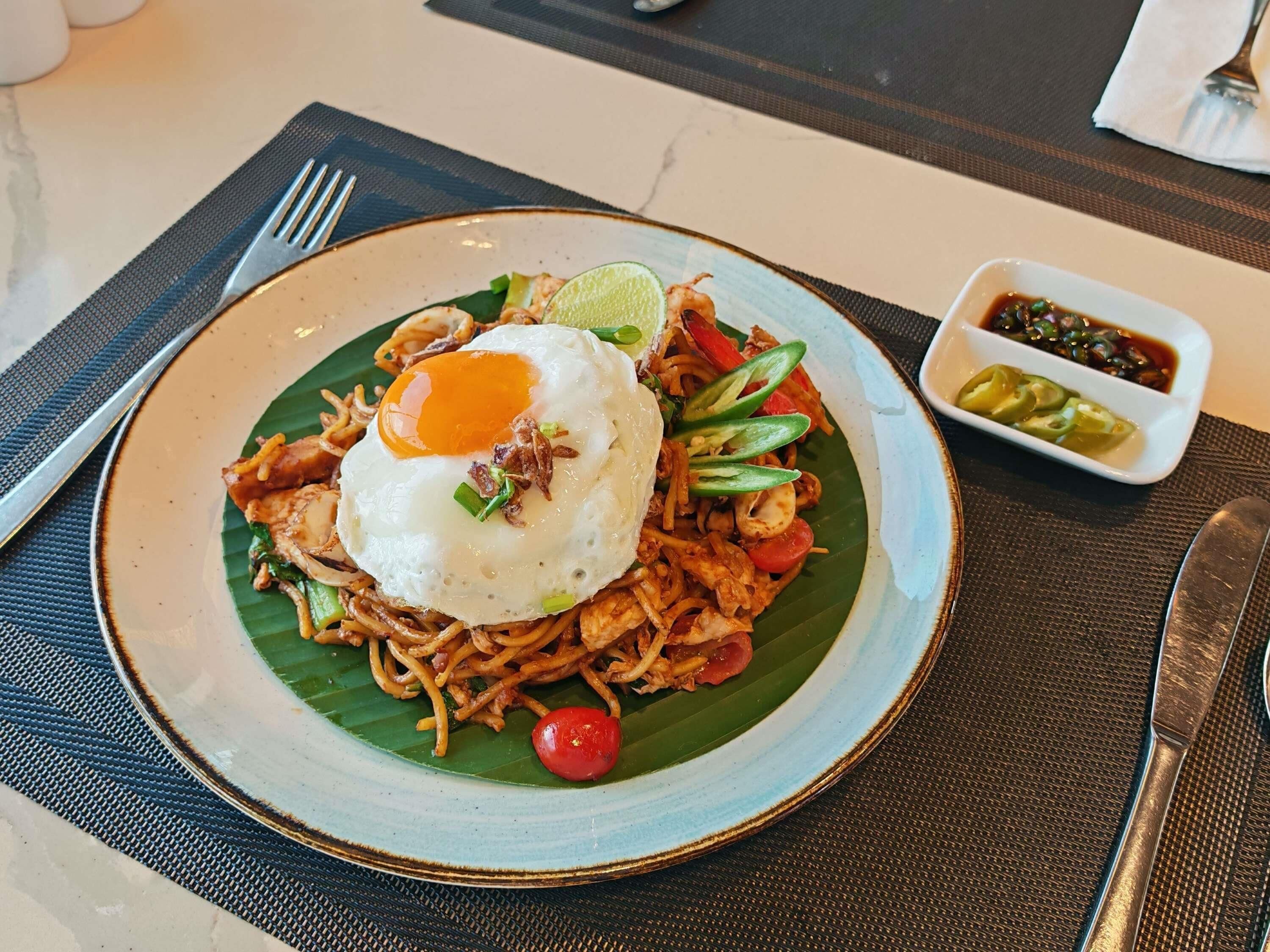 A plate of noodles with a fried egg, garnished with green onions and a lime wedge, served on a banana leaf.