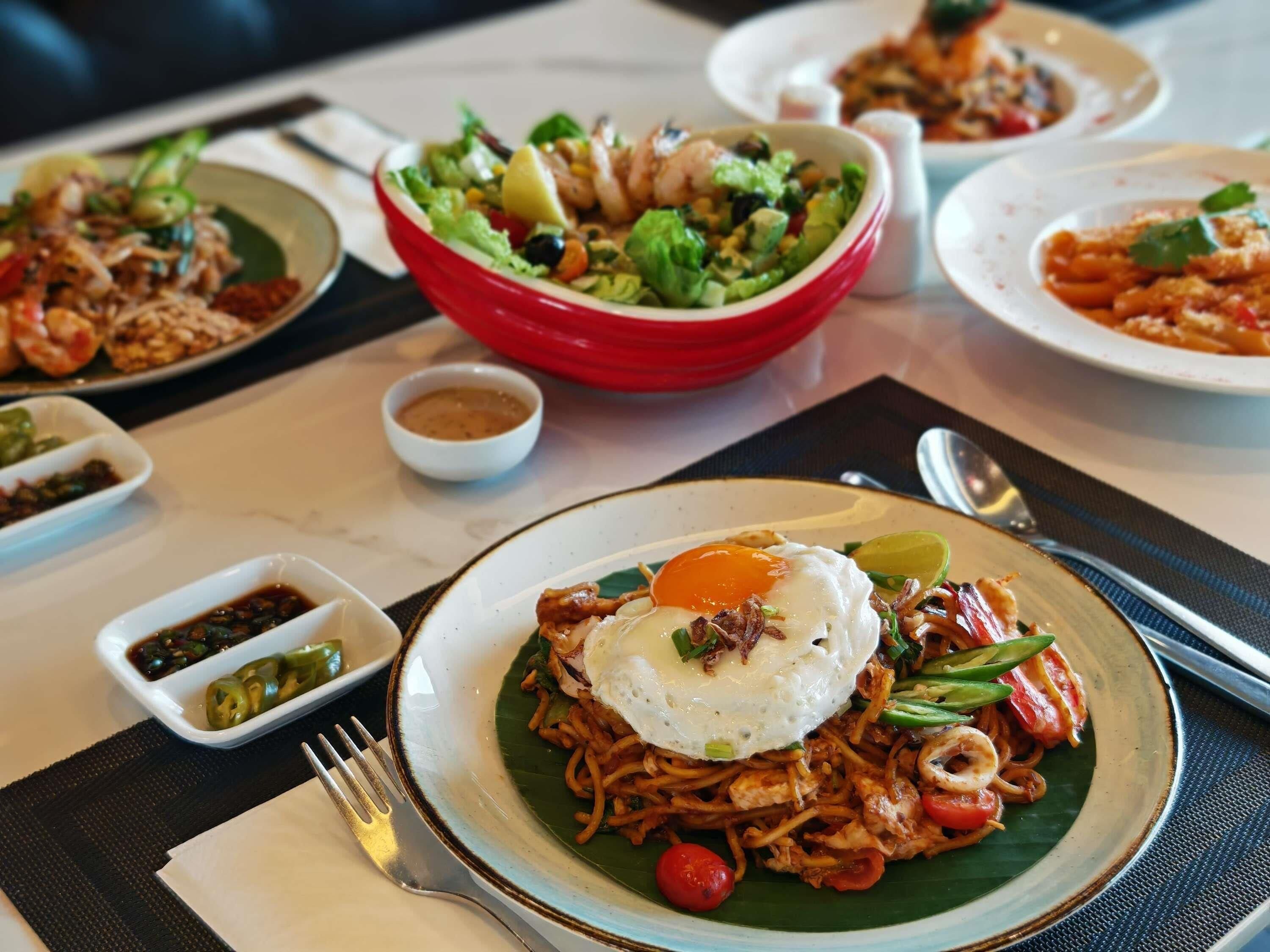 Une table où l’on trouve différents plats, notamment des nouilles aux œufs, aux crevettes et aux légumes, ainsi qu’un saladier rouge rempli de salade.