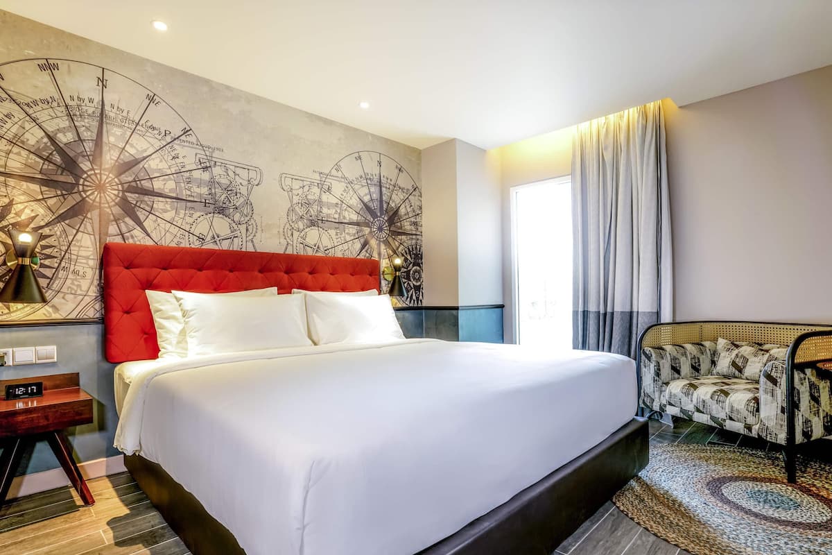 Hotelkamer met een groot bed, een rode hoofdbord, een wandklok, een nachtkastje, een gestreept fauteuil en een grote wandkaart.