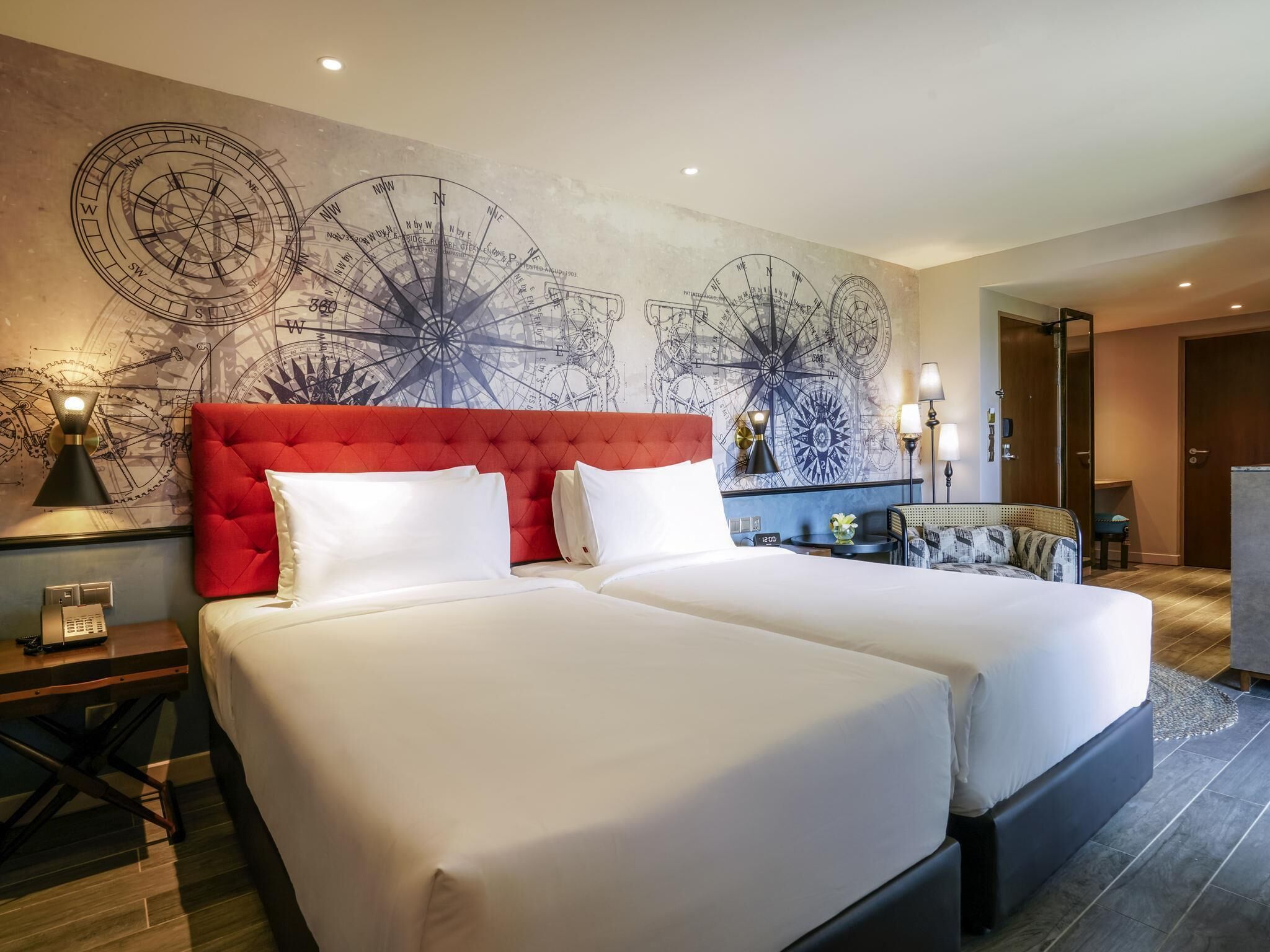A hotel room with two beds, a red headboard, a large wall map, a desk with a lamp, and a small table with a vase.