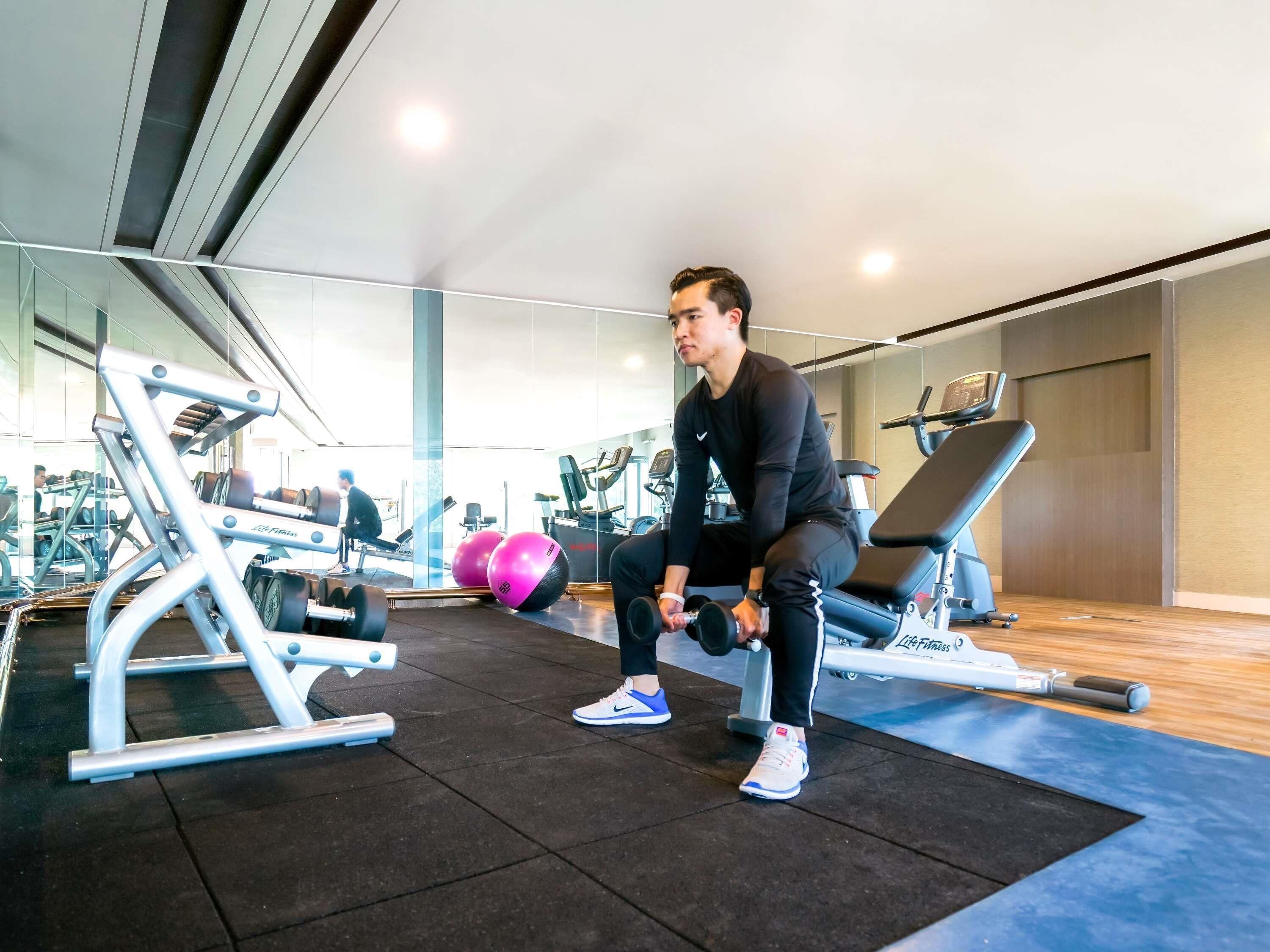 A gym with exercise equipment, a mirrored wall, and a person using dumbbells.