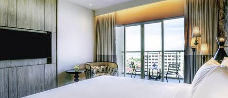 A hotel room with a large bed, a TV mounted on the wall, a small table with a flower, and a balcony with chairs and a table.