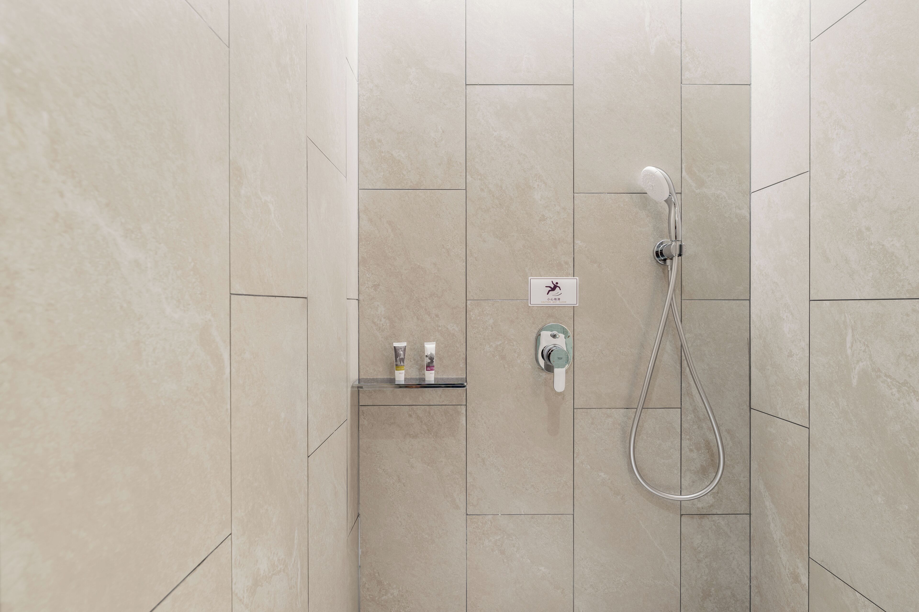 Área de duche com chuveiro manual, painel de controlo fixado na parede e prateleira com artigos de higiene pessoal.
