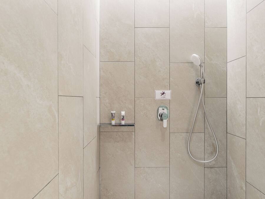 Área de duche com chuveiro manual, painel de controlo fixado na parede e prateleira com artigos de higiene pessoal.