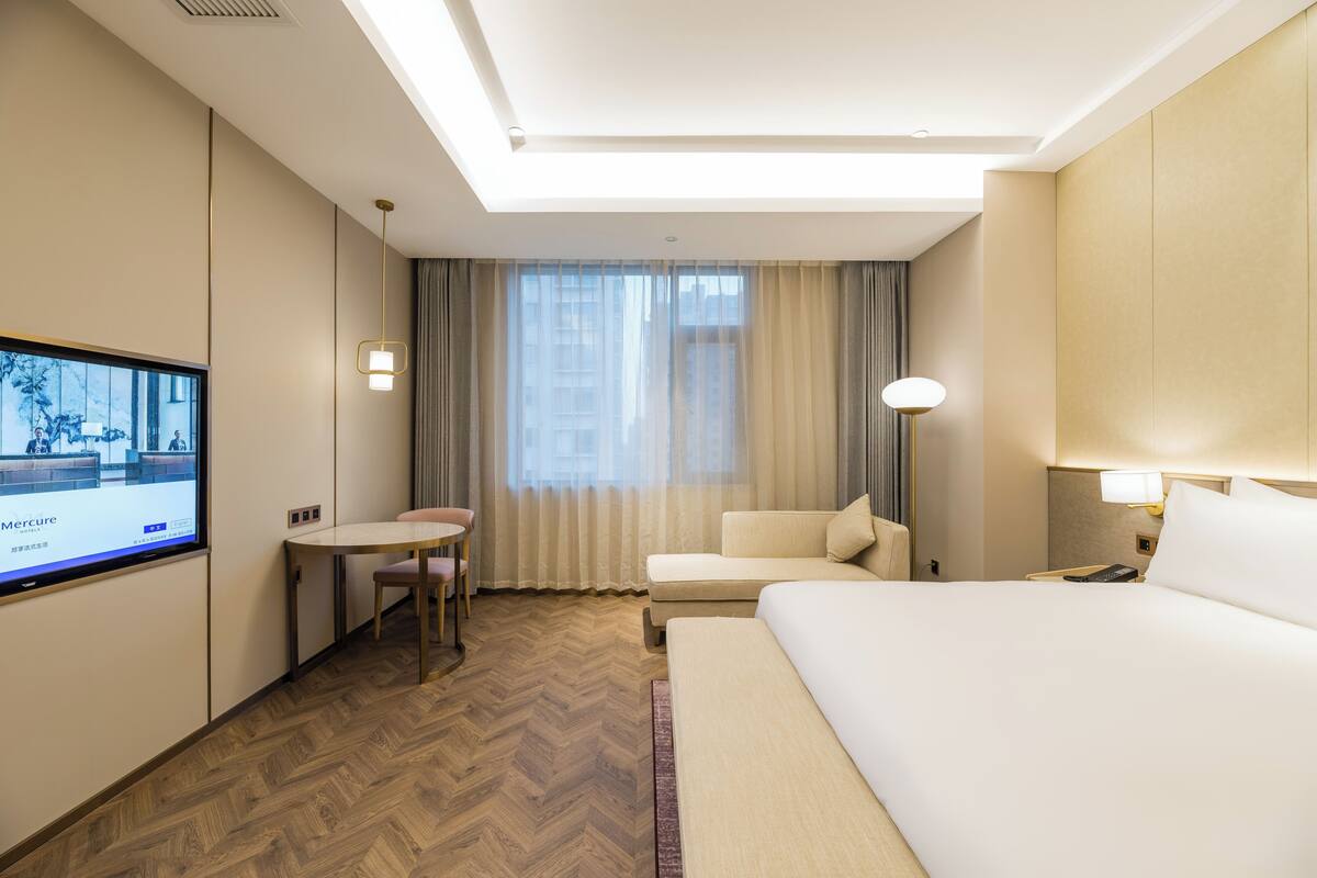 Quarto de hotel com cama, escrivaninha, cadeira, televisão e janela com cortinas.
