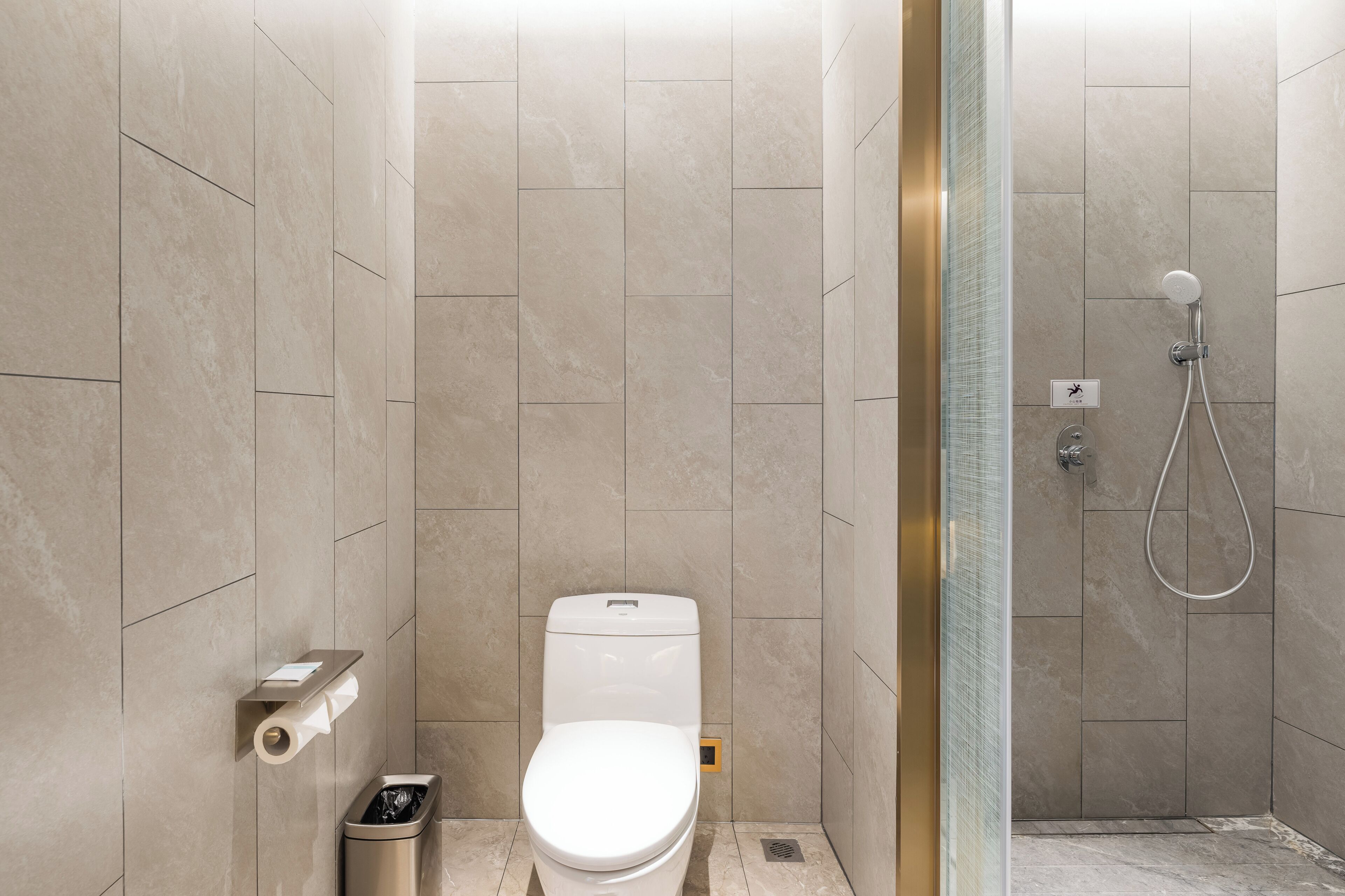 A bathroom with a toilet, a trash bin, a shower with a glass door, and a wall-mounted toilet paper holder.
