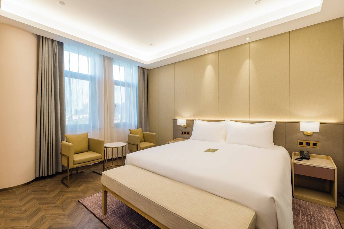 Quarto de hotel com uma cama grande, mesas de cabeceira, uma pequena mesa redonda, duas poltronas e uma janela com cortinas.