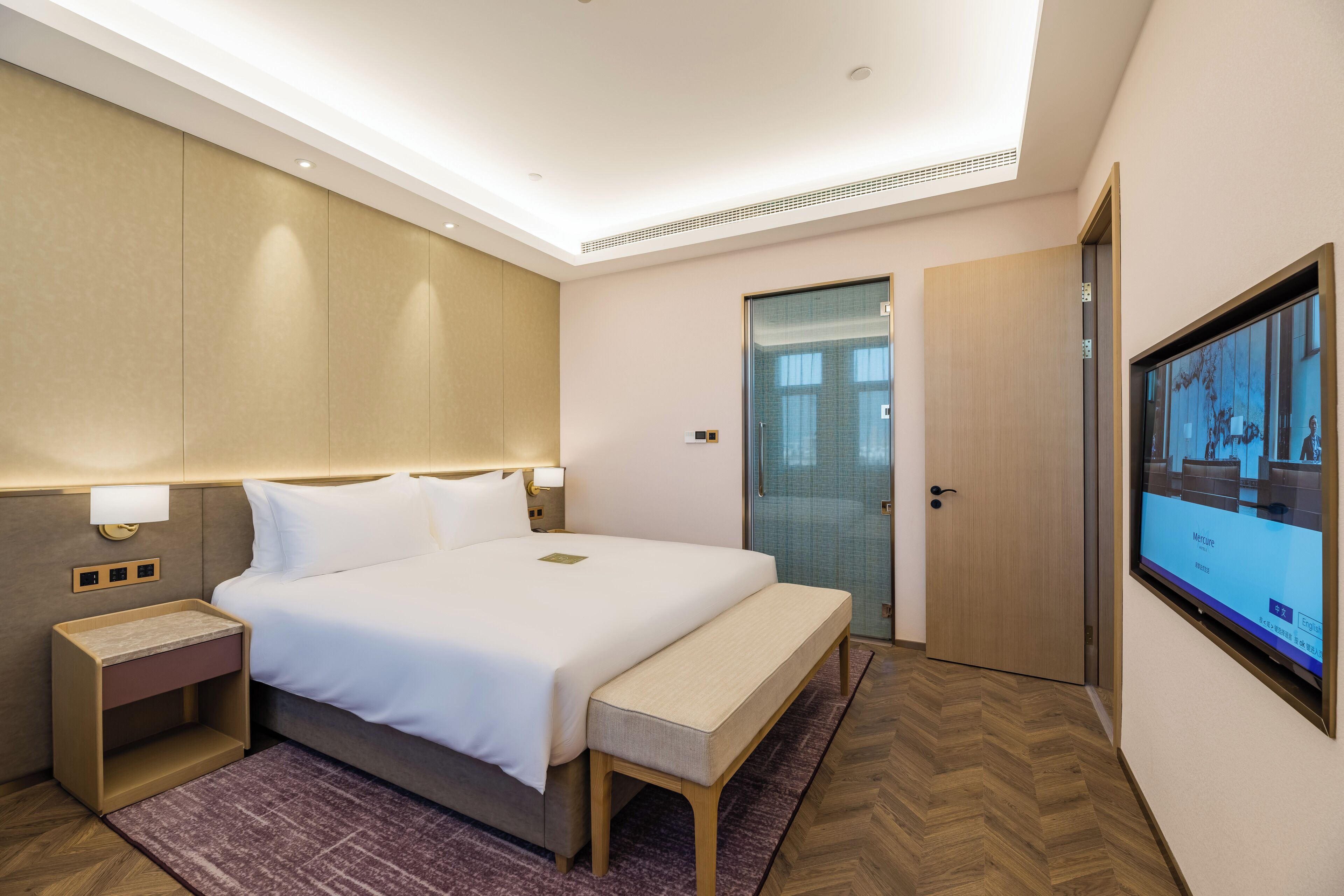 Habitación de hotel con una cama grande, mesitas de noche, un banco, un televisor y una puerta.