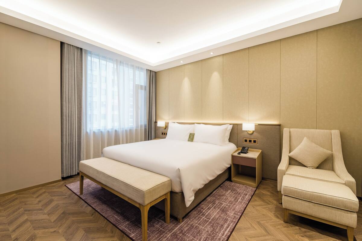Quarto de hotel com uma cama grande, mesa de cabeceira, cadeira, janela com cortinas e piso de madeira.
