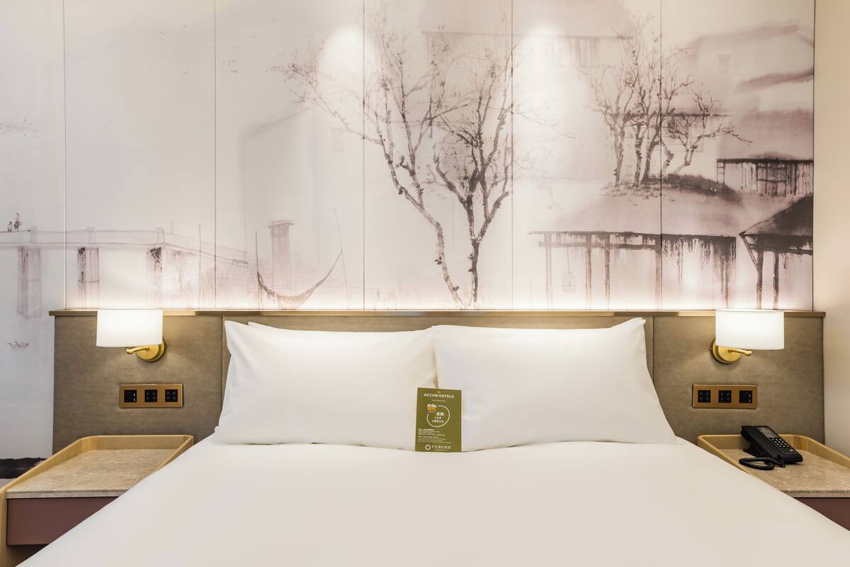 Quarto de hotel com cama, duas luminárias de cabeceira, mesa de cabeceira com telefone e uma obra de arte na parede.