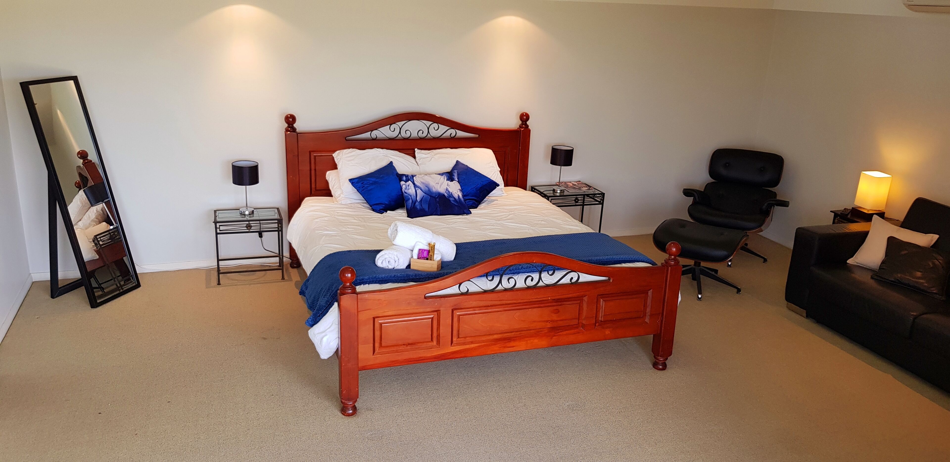 Una cama bien hecha con tablero de madera, dos mesitas de noche con lámparas, un sillón de cuero negro y un sofá de cuero negro.