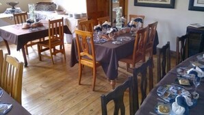 A dining area with wooden tables and chairs, framed artwork on the walls, and a window with curtains. - Rosenhof Country Lodge (Paul Roux)