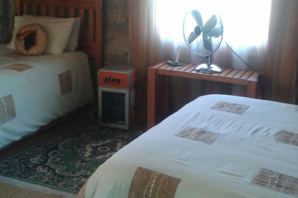 Ein Hotelzimmer mit zwei Betten, einem kleinen Tisch mit Ventilator, einer Spielstation und einem großen Fenster mit Vorhängen.