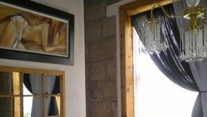 A rustic bathroom with a stone wall, a wooden window with curtains, a black toilet, and a framed artwork above the window. - Rosenhof Country Lodge (Paul Roux)