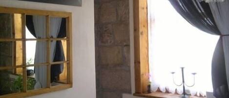 Un baño rústico con pared de piedra, ventana de madera con cortinas, inodoro negro y una obra de arte enmarcada sobre la ventana.