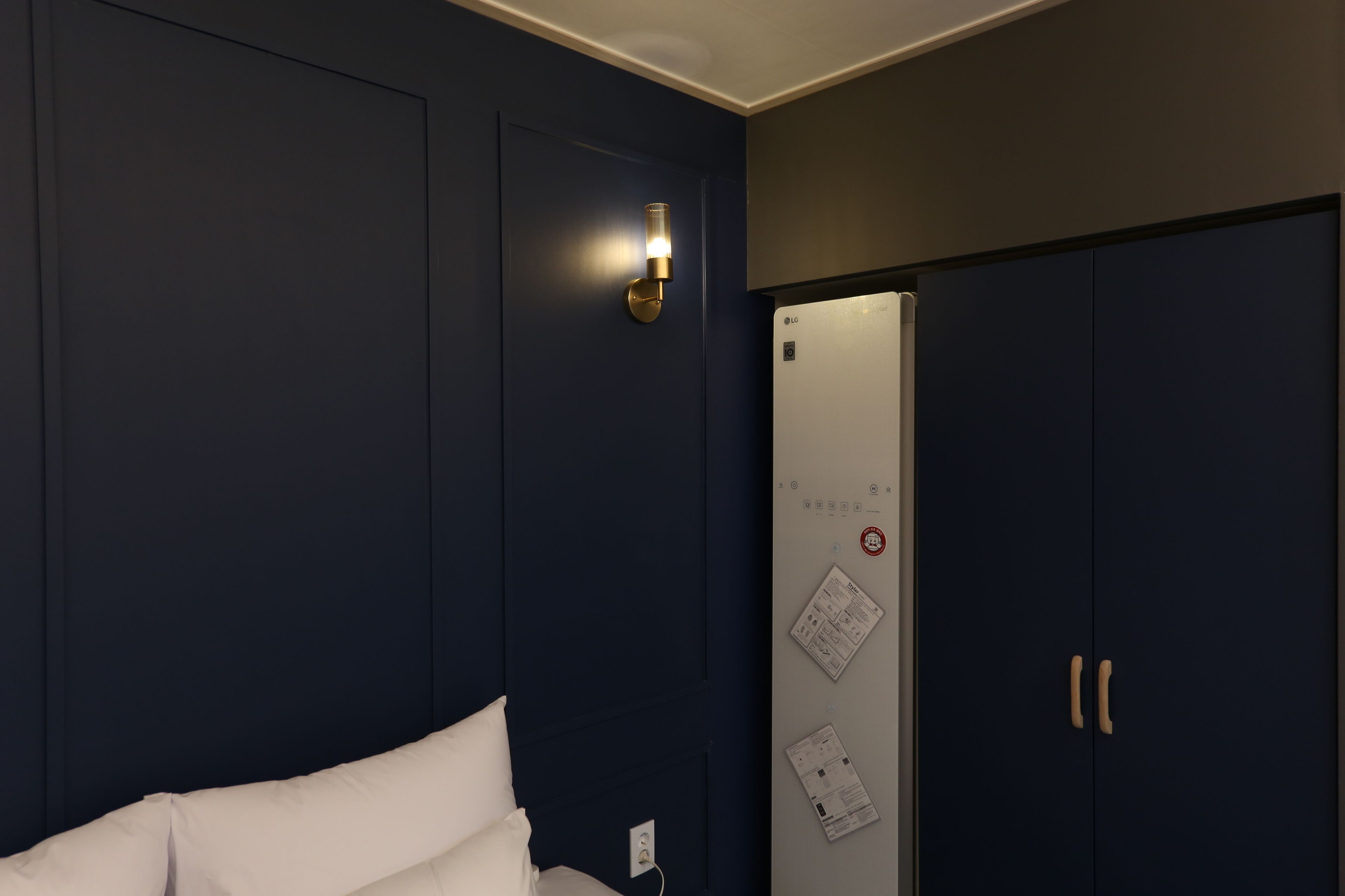 A hotel room with a bed, a wall-mounted light fixture, a refrigerator, and a door with a lock.