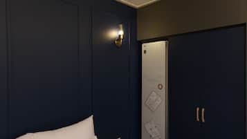 A hotel room with a bed, a wall-mounted light fixture, a refrigerator, and a door with a lock.
