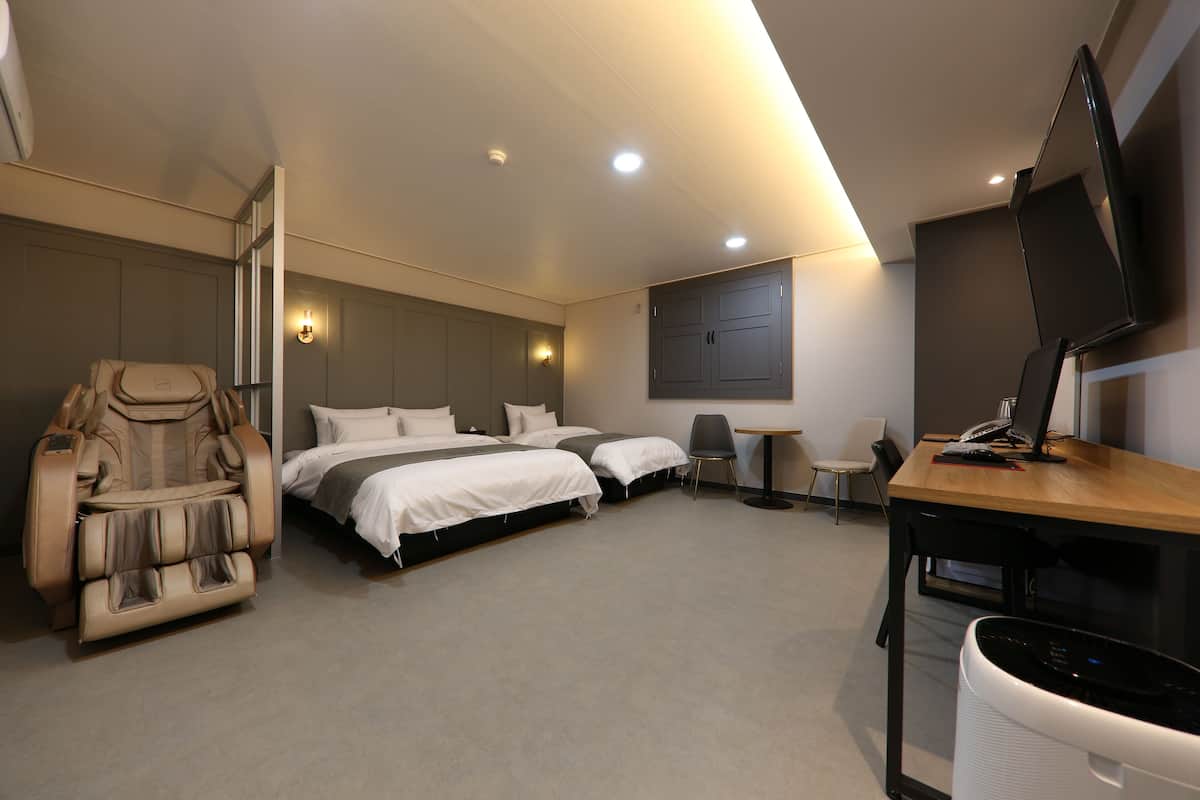 Una habitación de hotel moderna con una silla de masajes, dos camas, un escritorio con una computadora y un televisor.