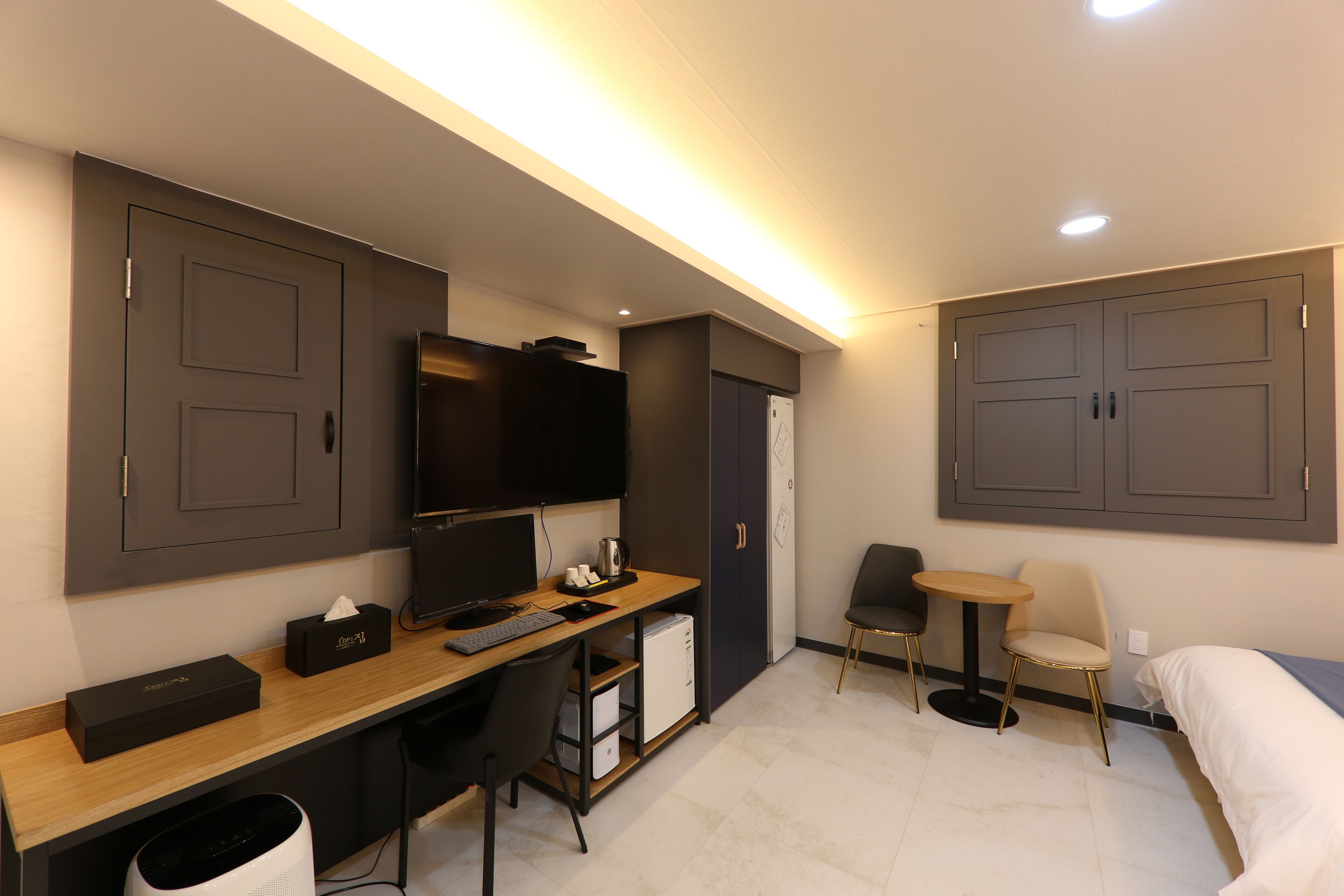 A modern hotel room with a wooden desk, a television, a small refrigerator, a chair, a bed, and a wardrobe.