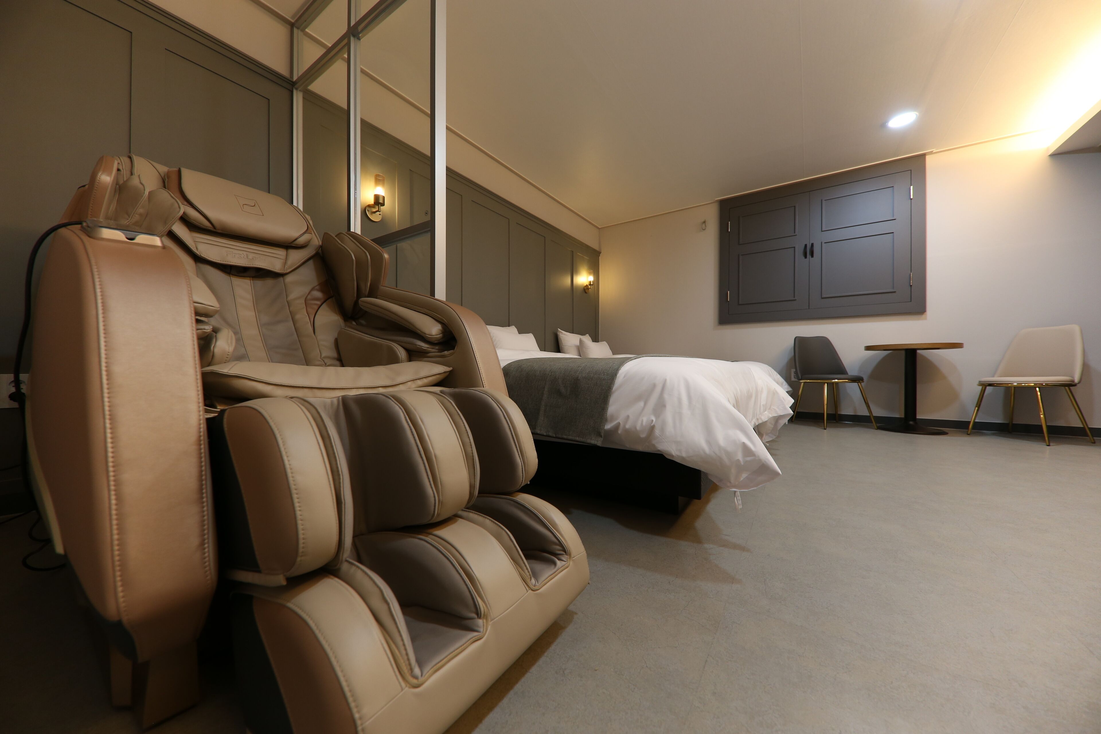A modern hotel room with a massage chair, a bed, a small table, and two chairs.