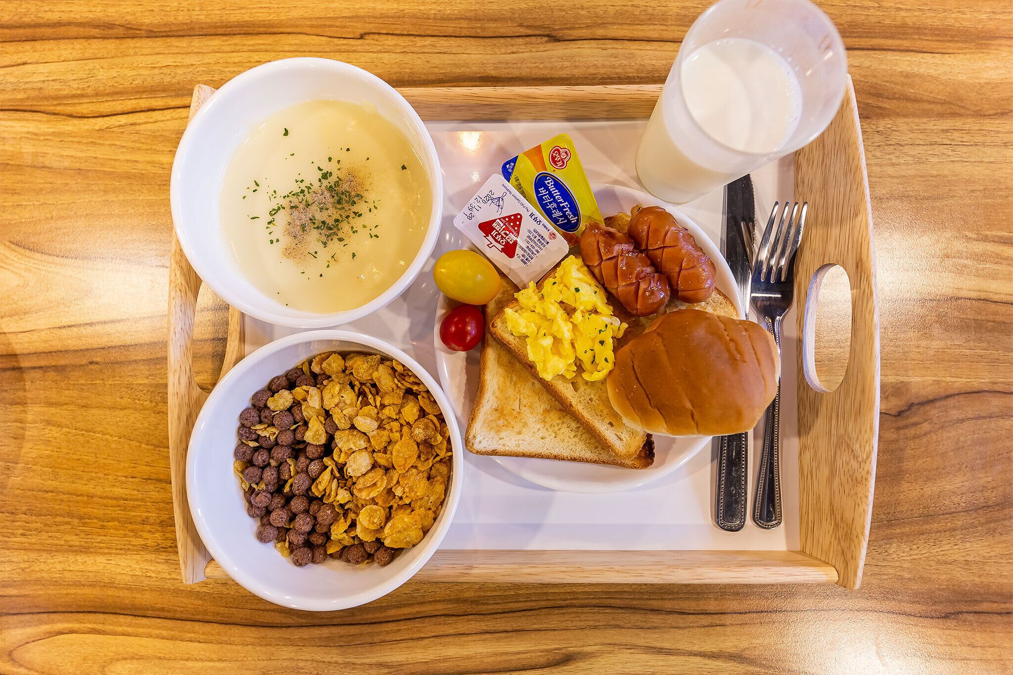 Una bandeja con un tazón de sopa, un plato con tostadas y huevos revueltos, un tazón de cereal, un vaso de leche y una cajita de mantequilla.