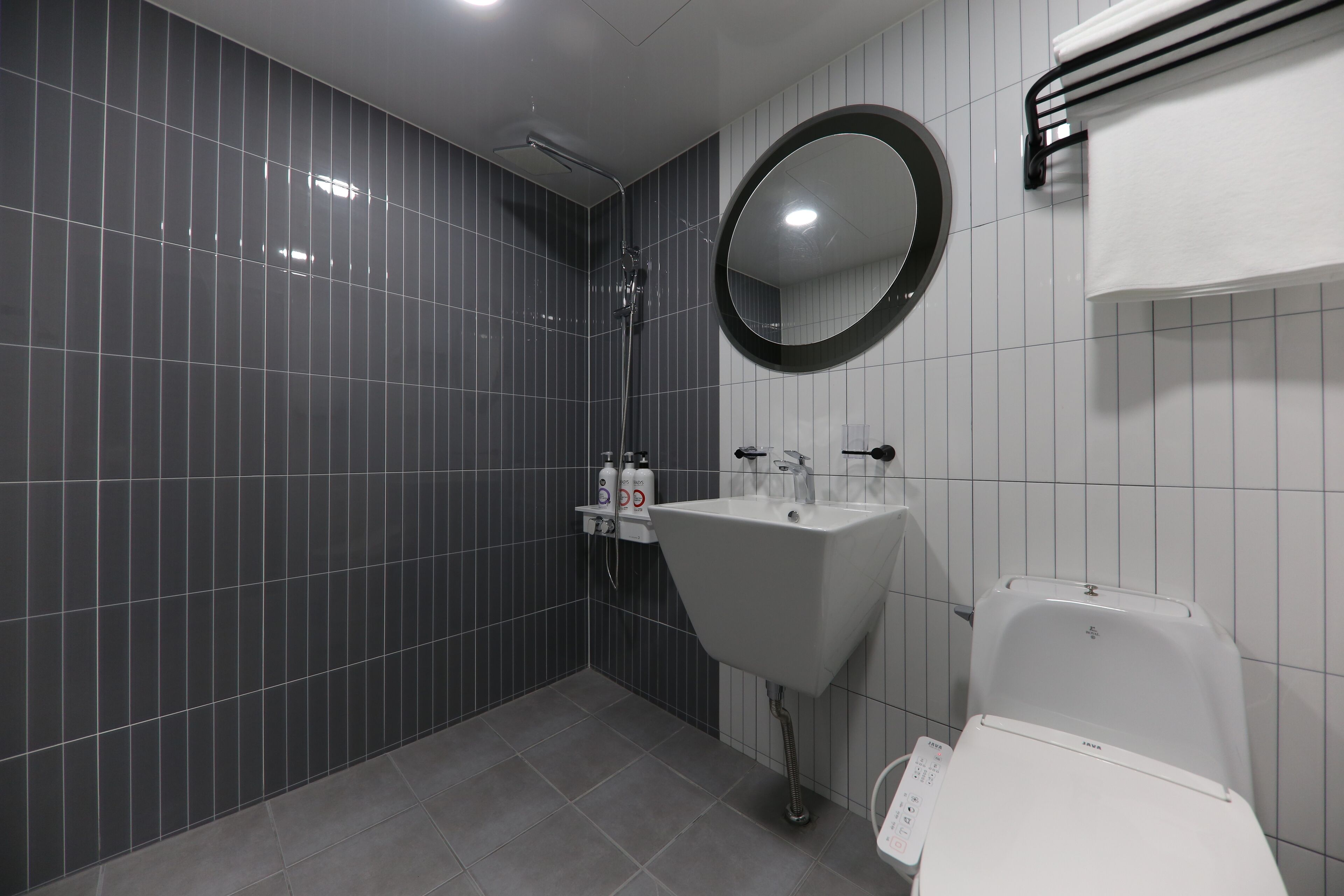 A modern bathroom with a grey tiled wall, a white sink, a round mirror, and a toilet.