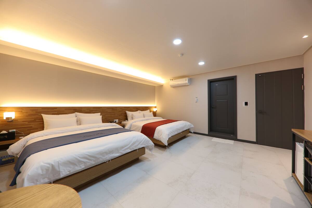 Habitación de hotel con dos camas, cabecero de madera, mesita de noche y un pequeño estante de madera, además de un equipo de aire acondicionado y una puerta.