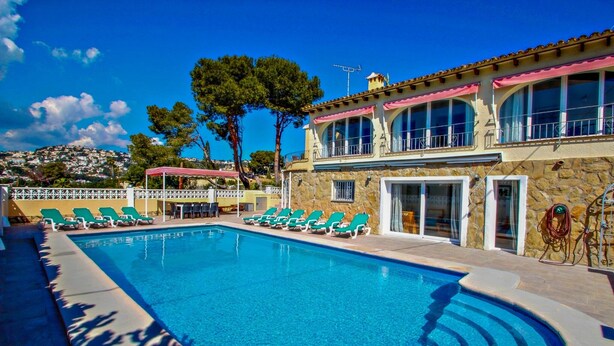Une piscine avec des chaises longues, un bâtiment en pierre doté de grandes fenêtres et une vue sur une colline.