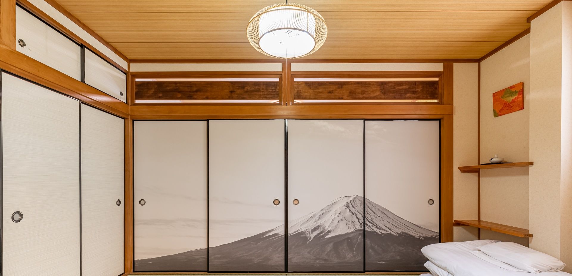 A room with a tatami mat floor, a bed with white bedding, a large sliding door with a mountain painting, and a ceiling light fixture.