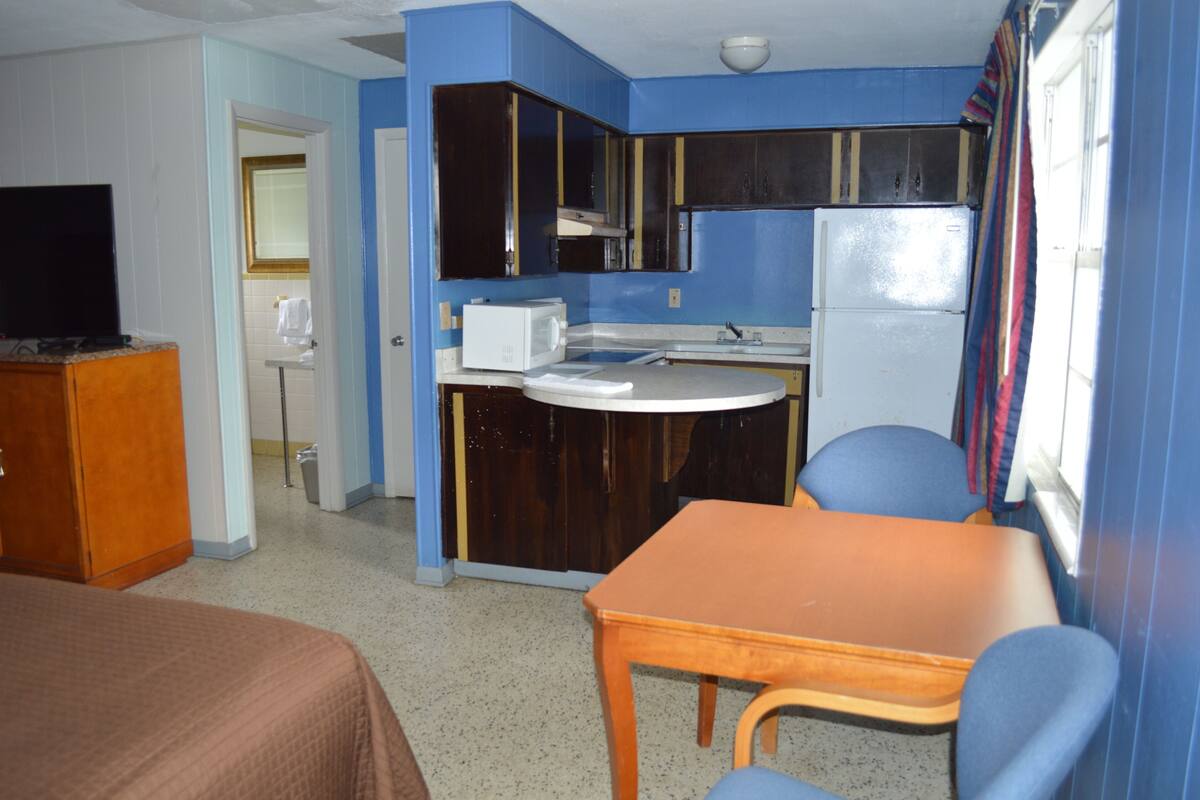 A compact hotel room with a kitchenette, a single bed, a small table, and a chair.