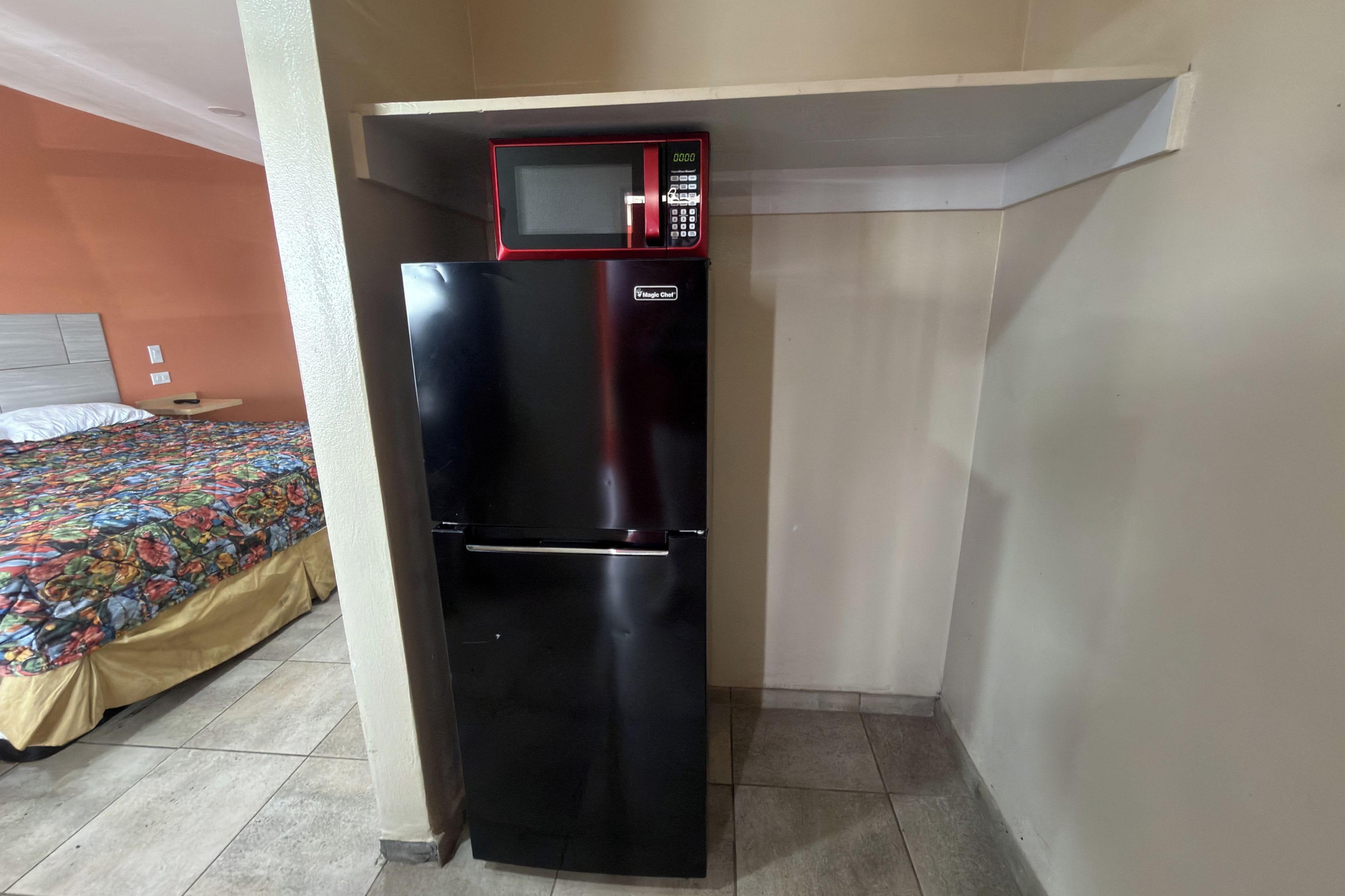 A compact black refrigerator with a microwave on top, positioned against a wall in a room with a bed and a colorful bedspread.