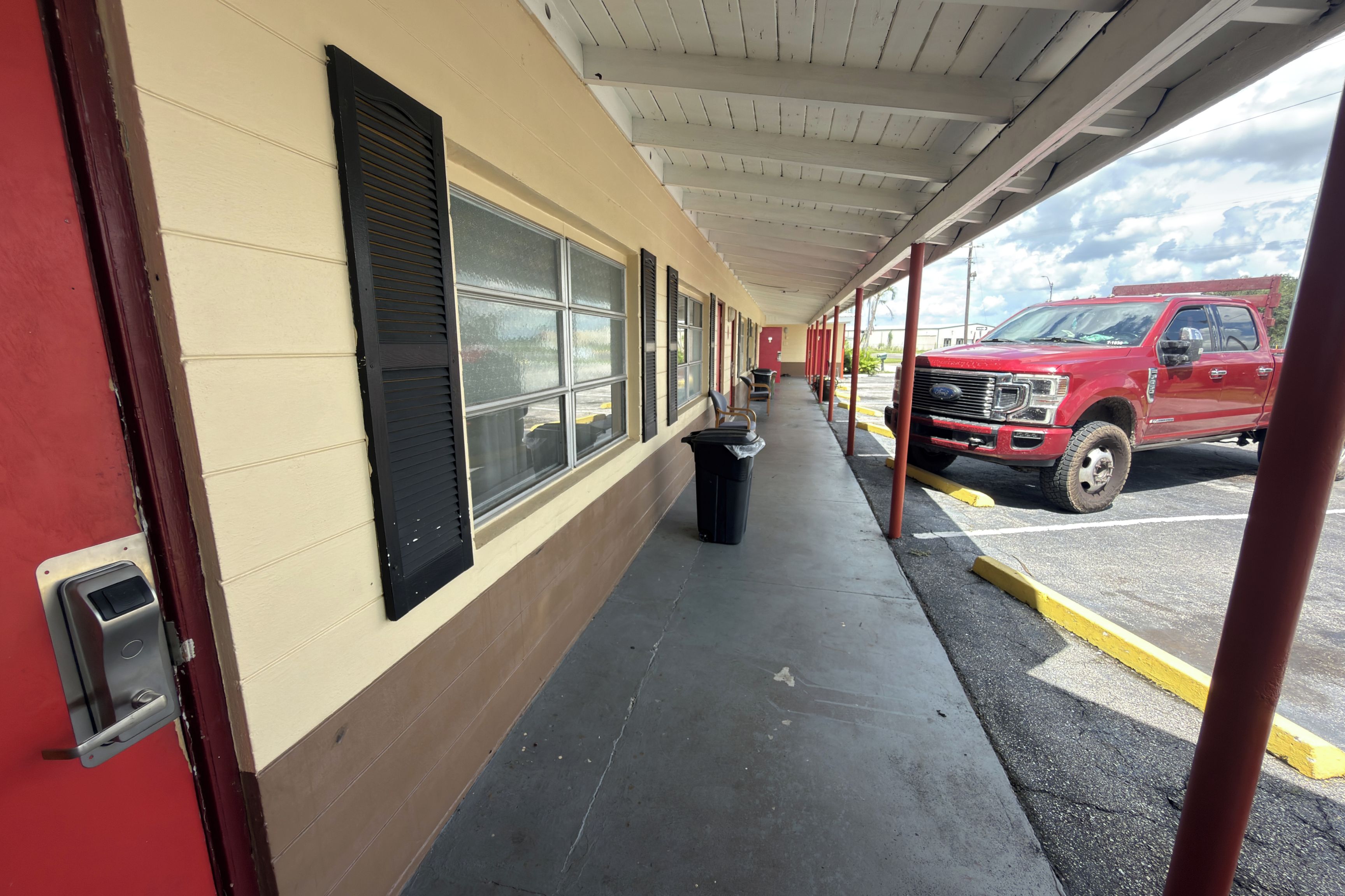 A covered corridor with a row of windows, a trash bin, and a red truck parked outside.