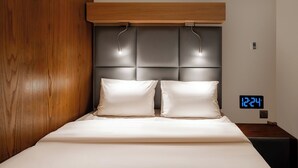Ein modernes Schlafzimmer mit einem Holz-Kopfteil, zwei weißen Kissen und einer digitalen Uhr, die 12:24 anzeigt.