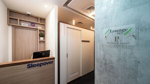 Ein Empfangsbereich mit einem Holztisch, der mit „Sleepover“ beschriftet ist, einem Schild mit der Aufschrift „Lounge Key Priority Pass“ und einer Überwachungskamera über dem Tisch.