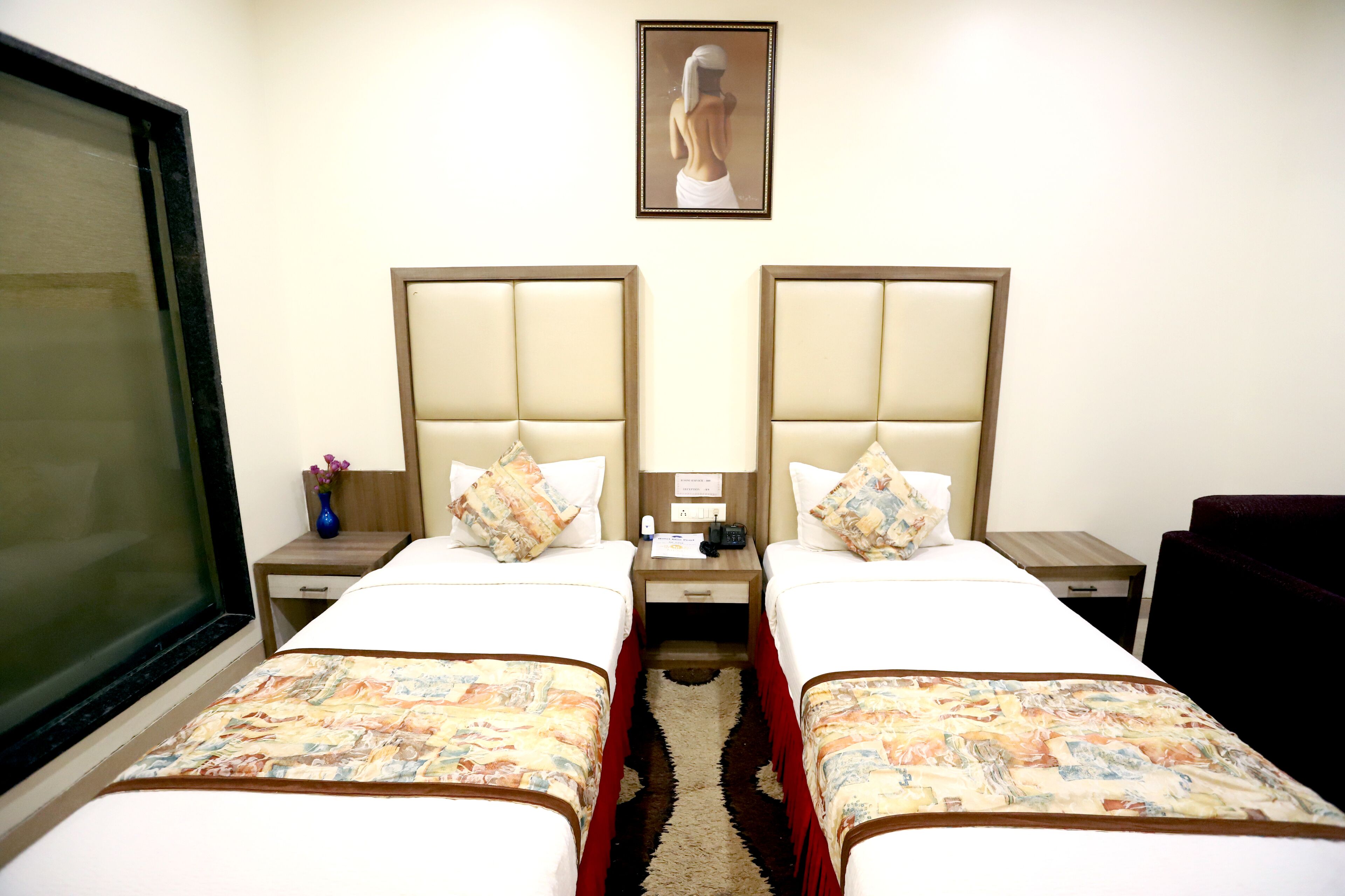 A hotel room with two beds, a nightstand with a lamp, a vase with flowers, and a framed picture on the wall.