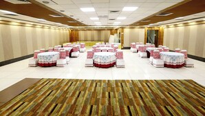 A large conference room with multiple round tables, each with a white tablecloth and red and white patterned chairs. The room has a tiled floor and a wooden panel wall. - HOTEL BLUE PEARL (Bhopal)