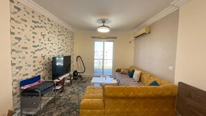 Una sala de estar con un sofá amarillo, un televisor, un aspirador y un tapete estampado.