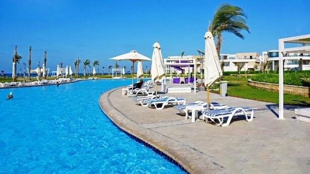 Un área de piscina con sillones de descanso y sombrillas, rodeada de palmeras y edificios.