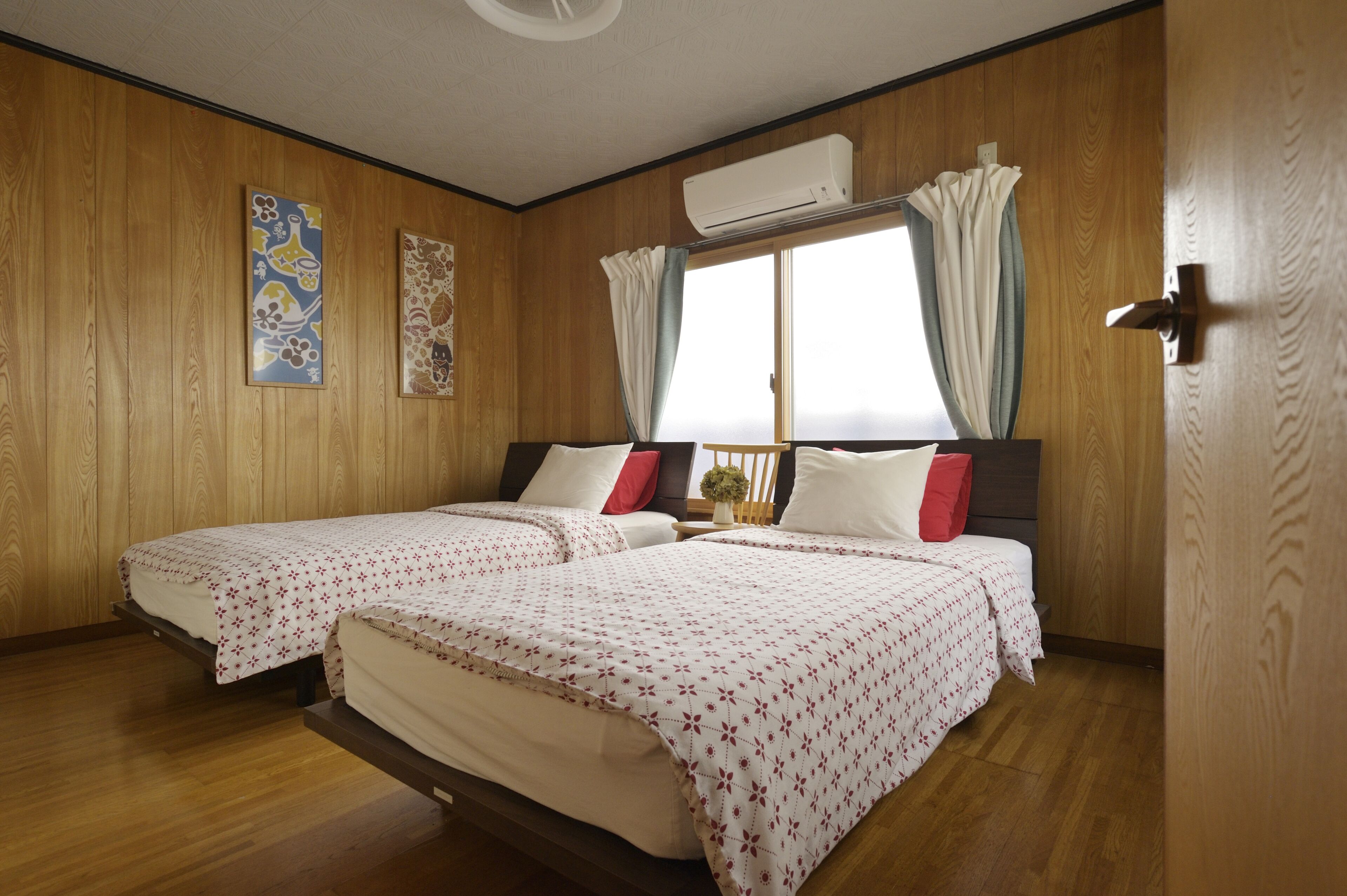 A room with two beds, wooden walls, an air conditioning unit, and a window with curtains.
