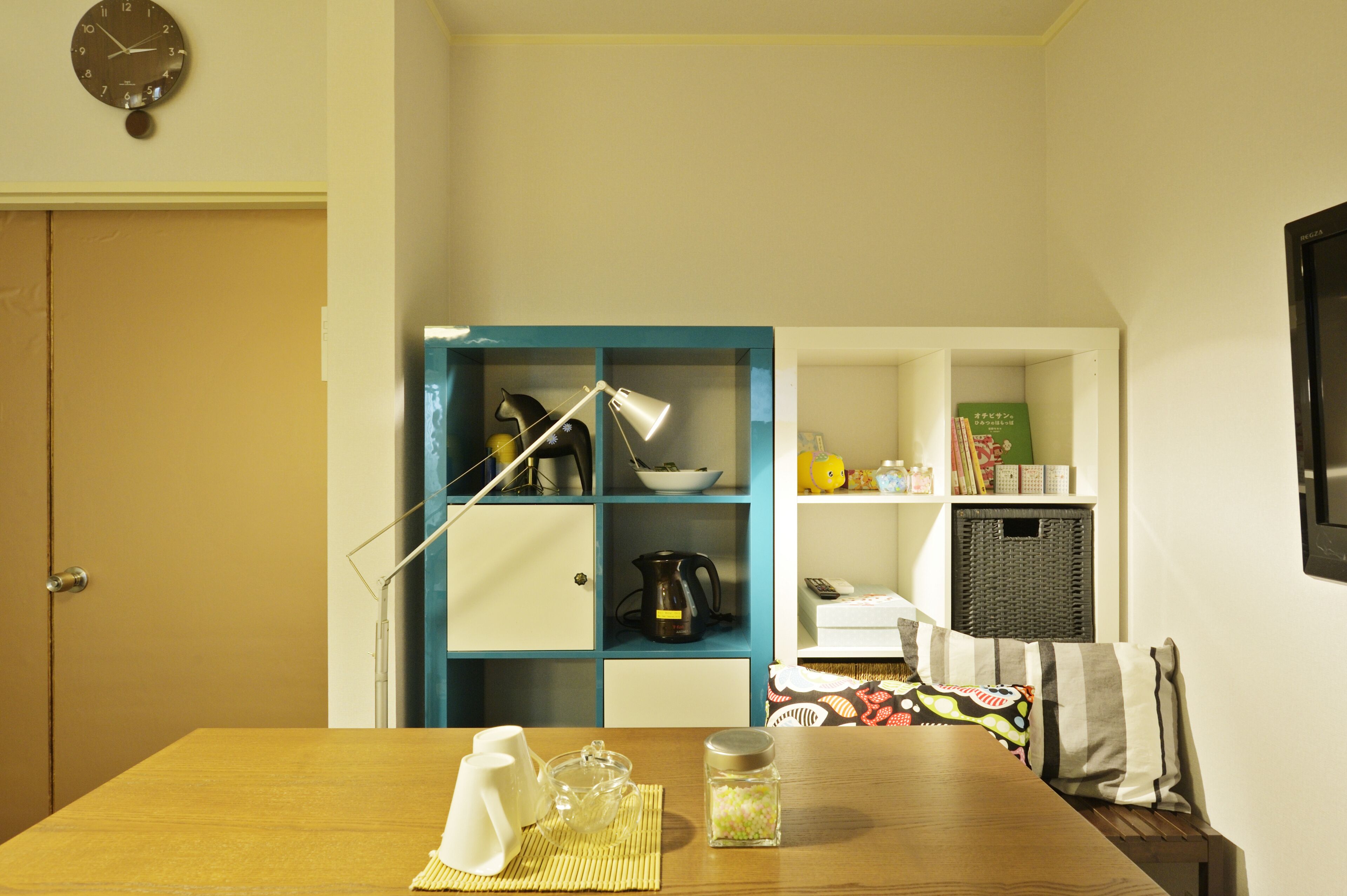 A room with a wooden table, a blue shelving unit with kitchen items, a clock on the wall, and a television on the right.